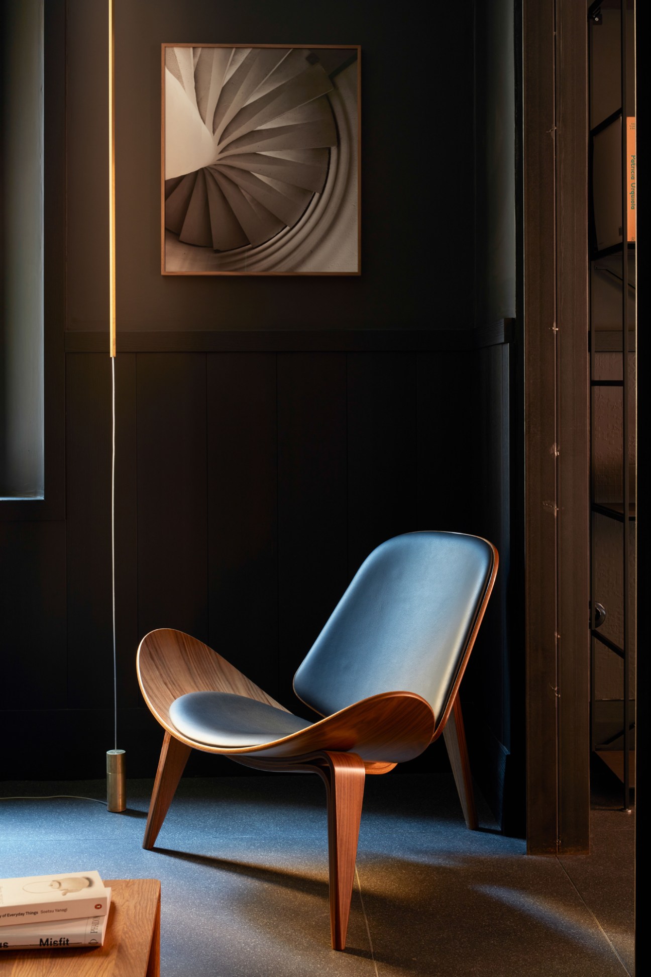 Modern minimalist interior with blue upholstered chair, wooden frame, and spiral staircase artwork on dark wall.