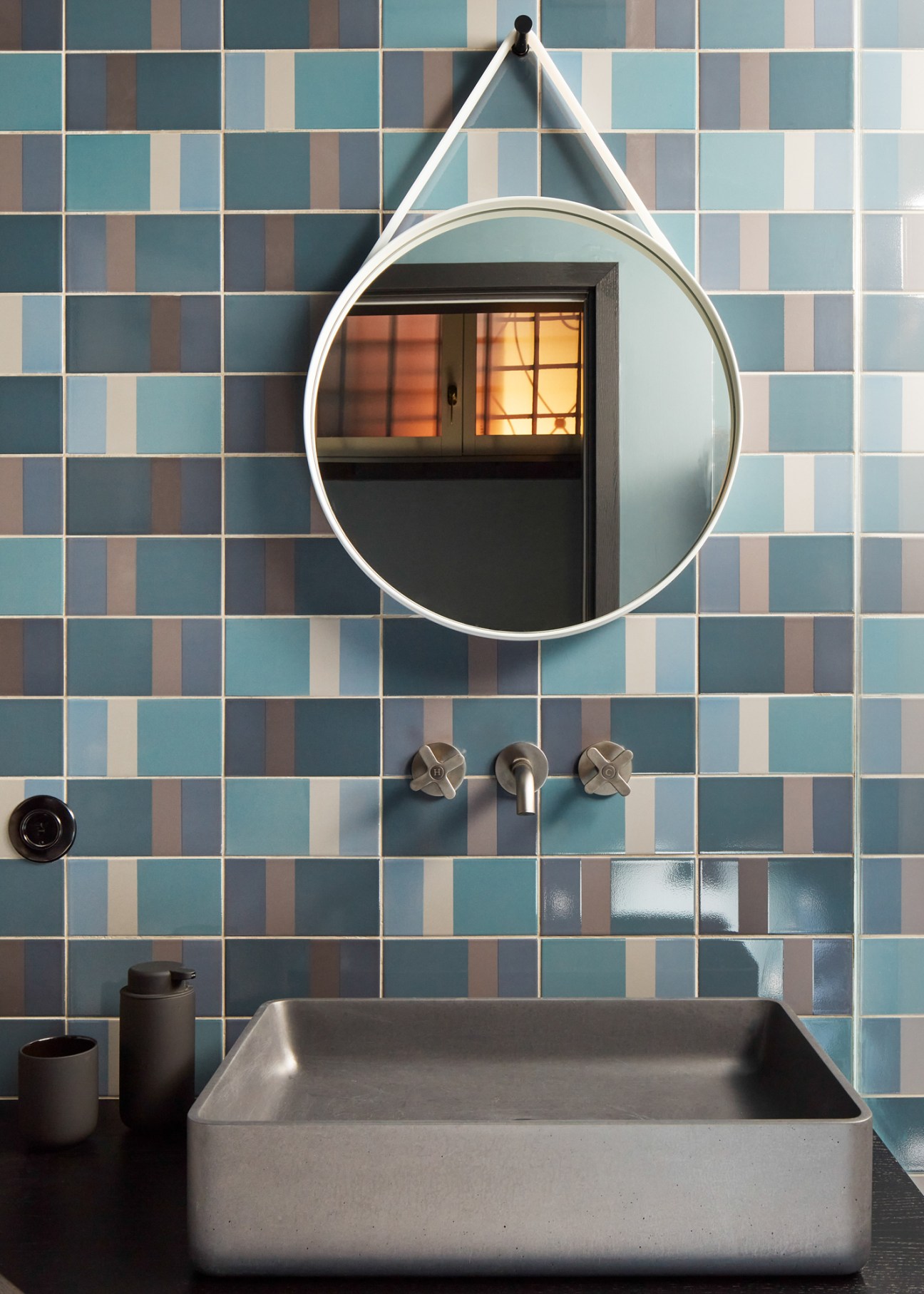 Bathroom with striped teal and beige tile wall, circular white mirror, concrete sink, and brass fixtures.