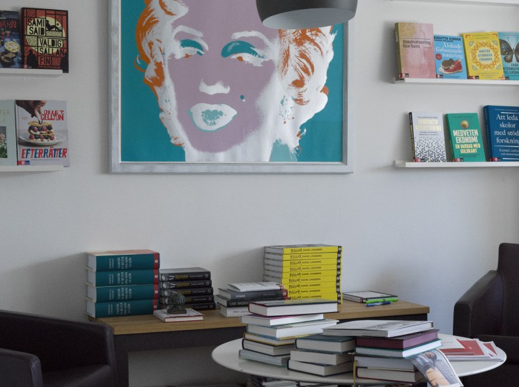 A reading lounge with stacked books on tables, wall-mounted shelves, and a large pop-art print above.