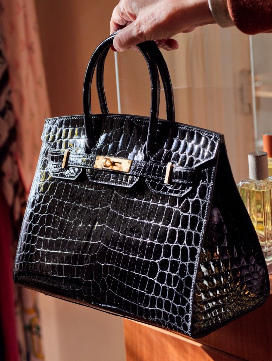 Hand holding a black crocodile leather handbag with gold hardware and turnlock closure in an elegant interior setting