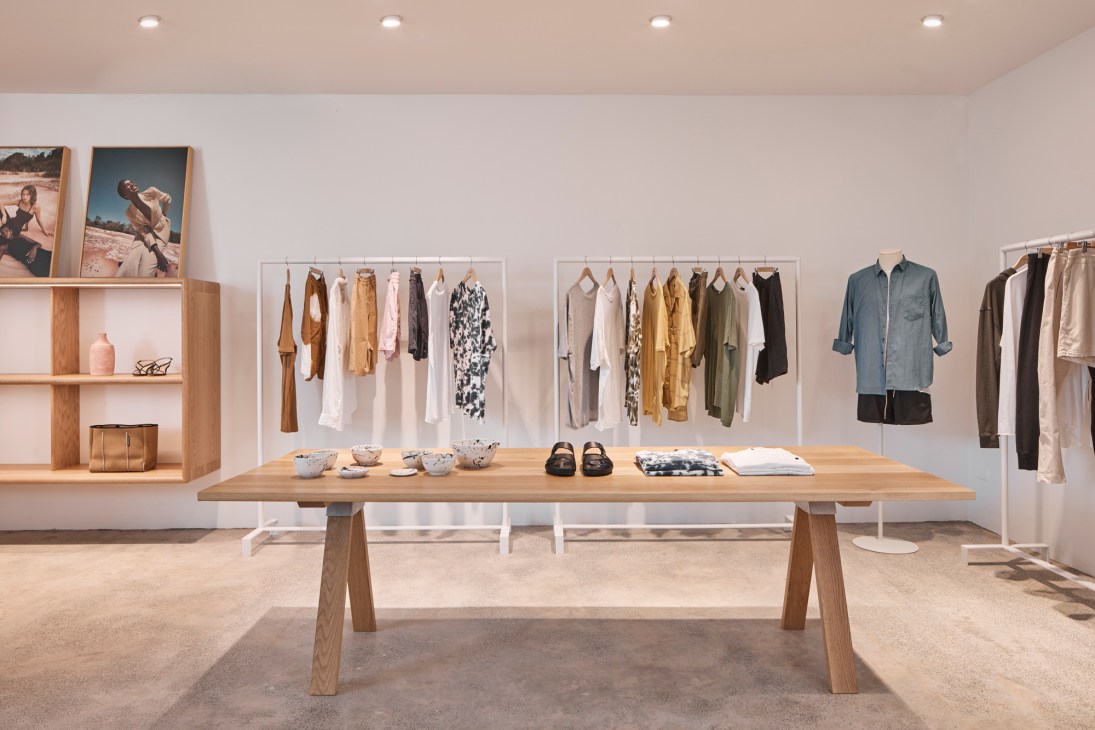 Interior of a minimalist clothing retail store with wooden display tables, neutral-toned walls, and racks of basic garments in earth tones.