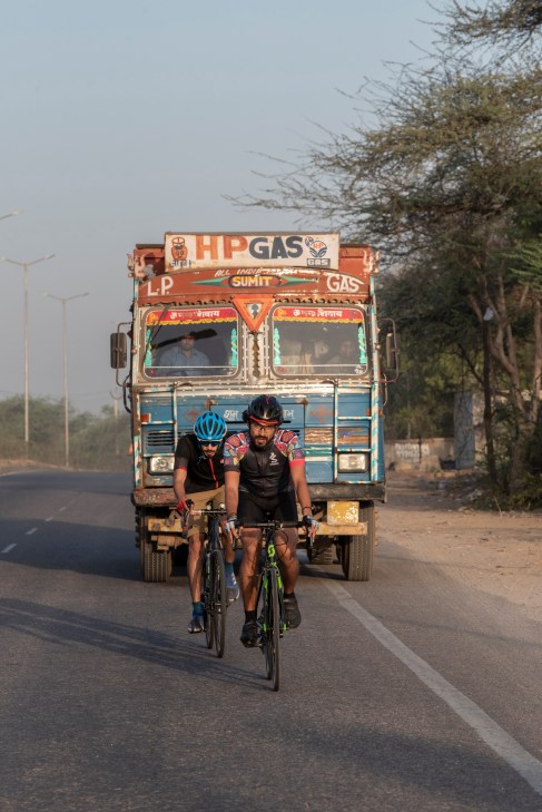 Two cyclists in helmets and athletic gear riding high-end bikes on a New Delhi road ahead of a colorful painted truck.