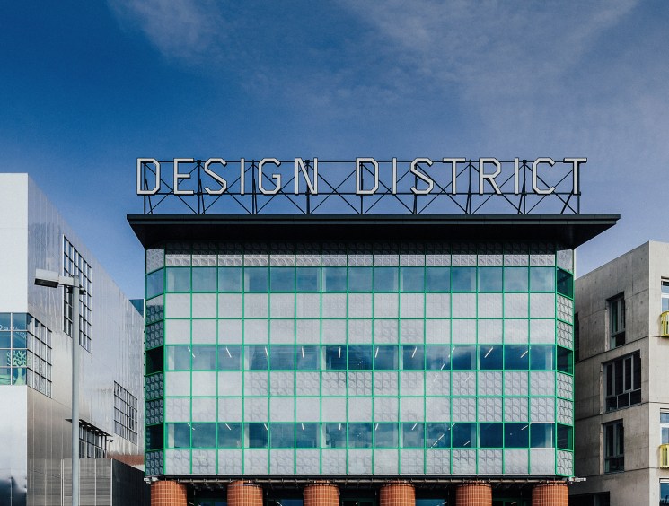 Modern building with green grid facade and 'DESIGN DISTRICT' sign on top surrounded by other contemporary structures