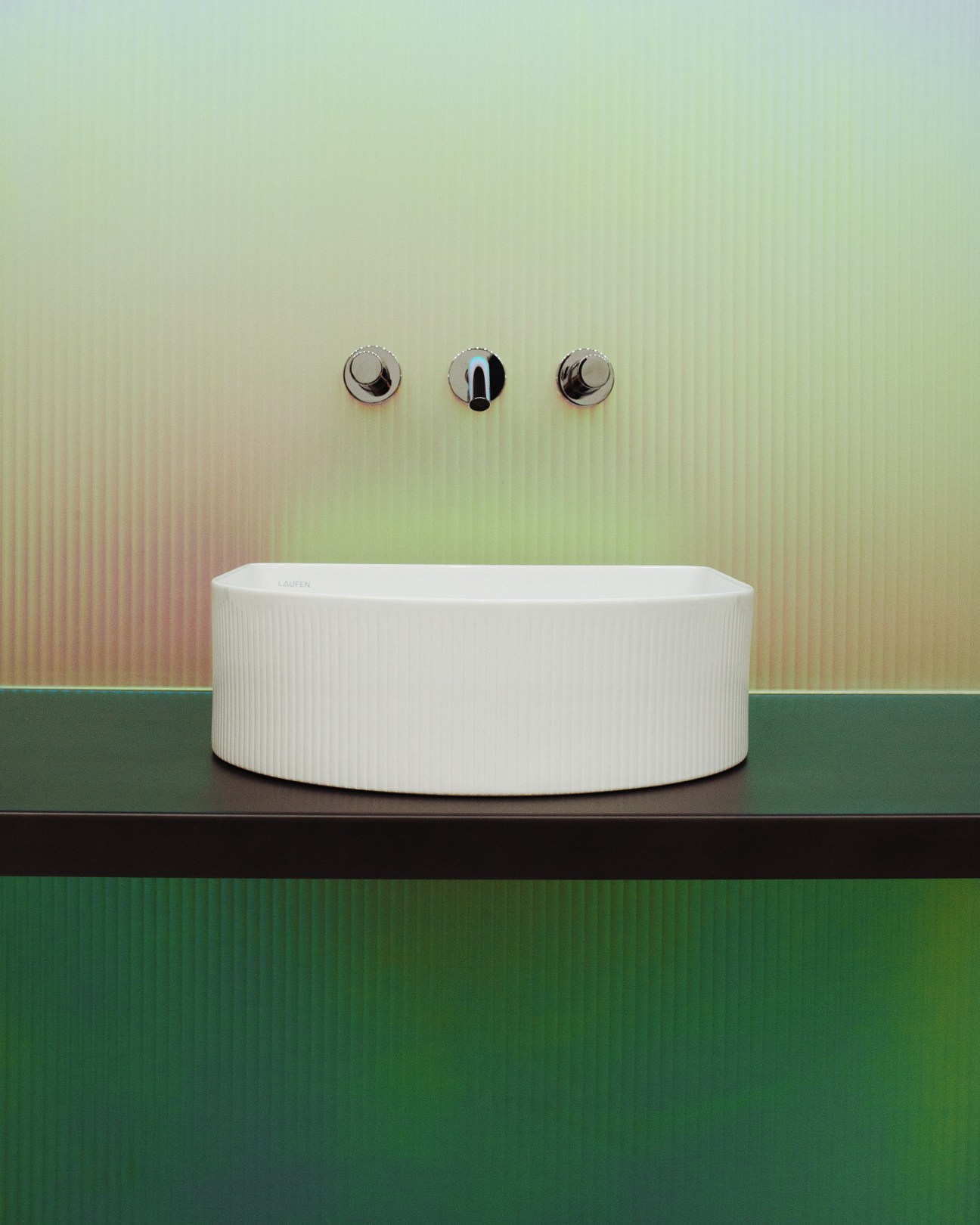 White ribbed basin by Patricia Urquiola for Laufen with three chrome wall-mounted faucet controls on a pastel green and pink gradient wall.
