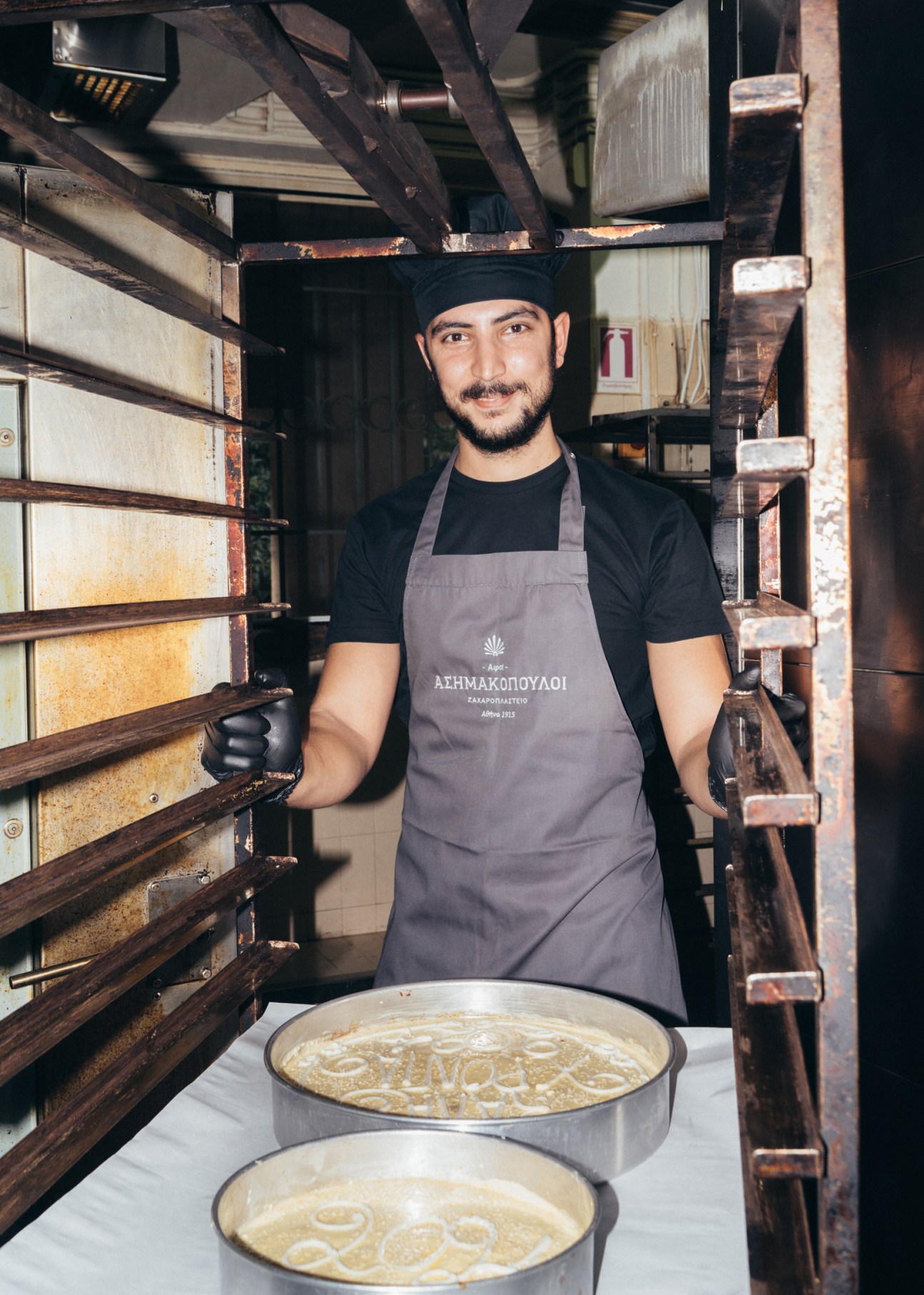 Baker in black beanie and gray apron stands smiling in traditional wooden bakery with fresh pastries on metal trays