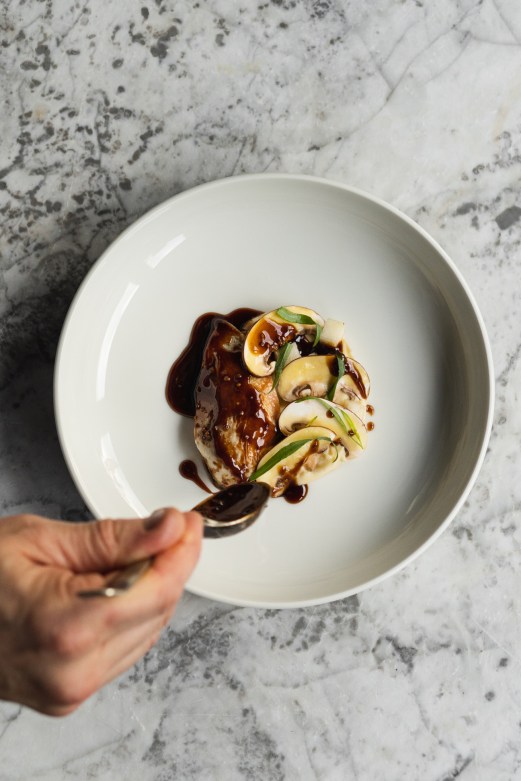 Plated dish with seared meat, mushrooms, apple slices and dark sauce on white ceramic plate held over marble counter.