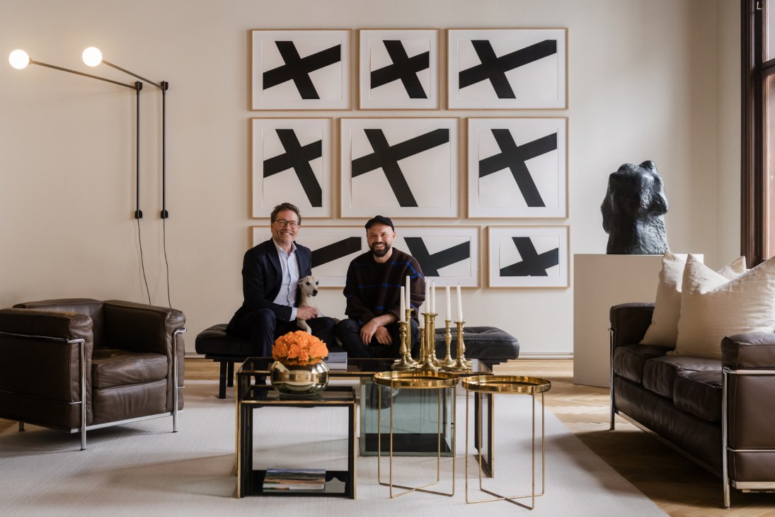 Two men seated on dark leather chairs in modern living room with nine framed black X artworks on wall