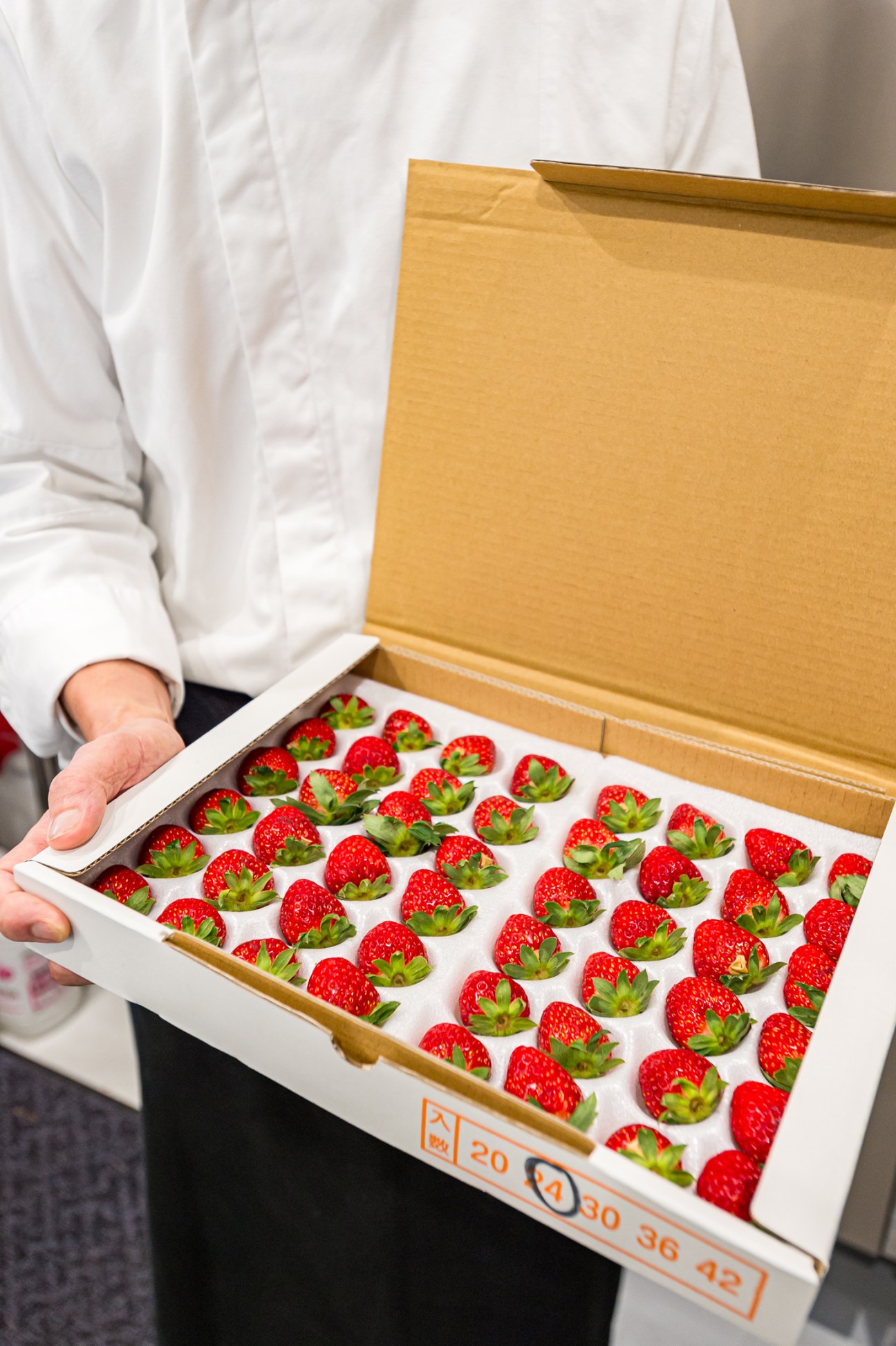 Person in white chef's coat holding an open box displaying rows of fresh red strawberries with green tops