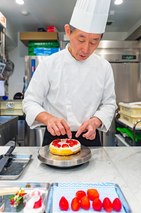 Chef in white uniform and hat carefully placing fresh strawberries on a small cream cake in professional kitchen
