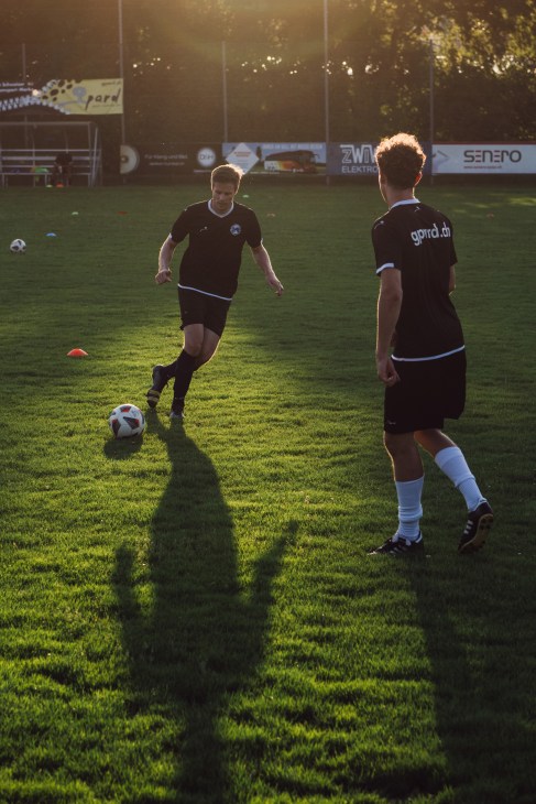 Two soccer players in dark uniforms practicing on a grass field during golden hour with stadium signage visible