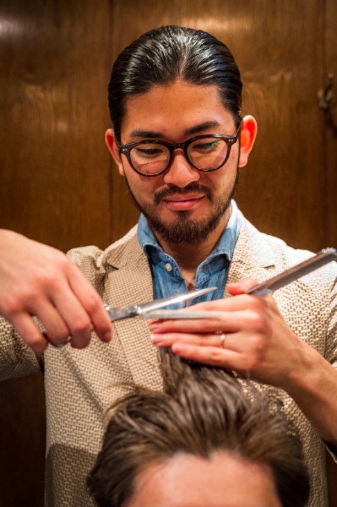 A barber wearing glasses and a beige cardigan carefully cuts a client's hair with scissors in a wooden-paneled salon