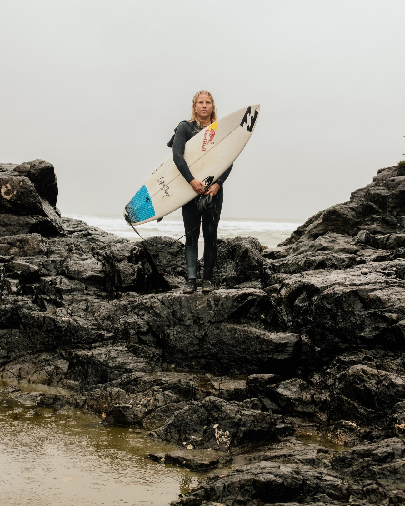 Blonde surfer in wetsuit holding white surfboard while standing on dark rocky coastline