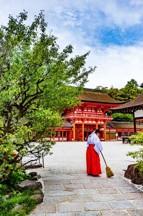 Japanese lady in white and red dress sweeping outside a red Japanese temple