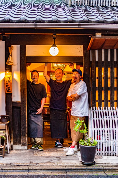 Three smiling men standing in doorway of traditional Japanese restaurant with warm lighting and tiled roof
