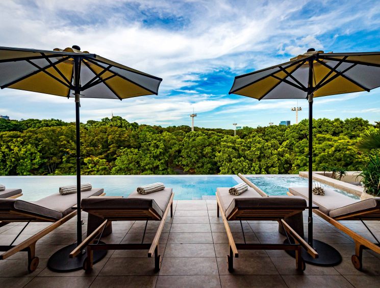 Pool deck with wooden loungers and umbrellas overlooking lush green forest canopy under blue sky