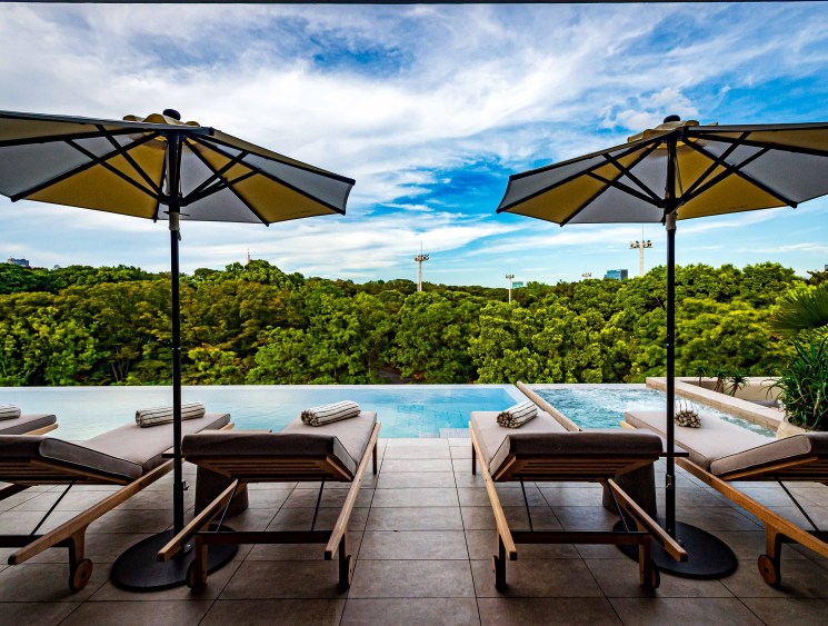 Pool deck with wooden loungers and umbrellas overlooking lush green forest canopy under blue sky