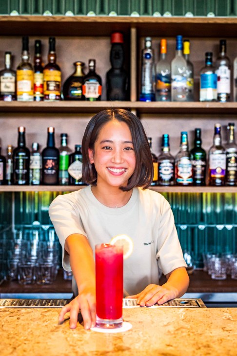 Smiling bartender in white shirt presents red cocktail with lemon garnish at modern bar with extensive bottle display