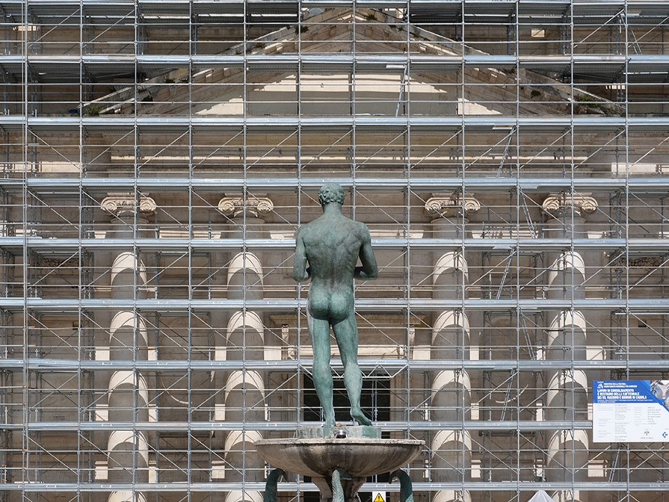 Bronze statue stands before scaffolding-covered historic Italian building during earthquake restoration work on Piazza Duomo