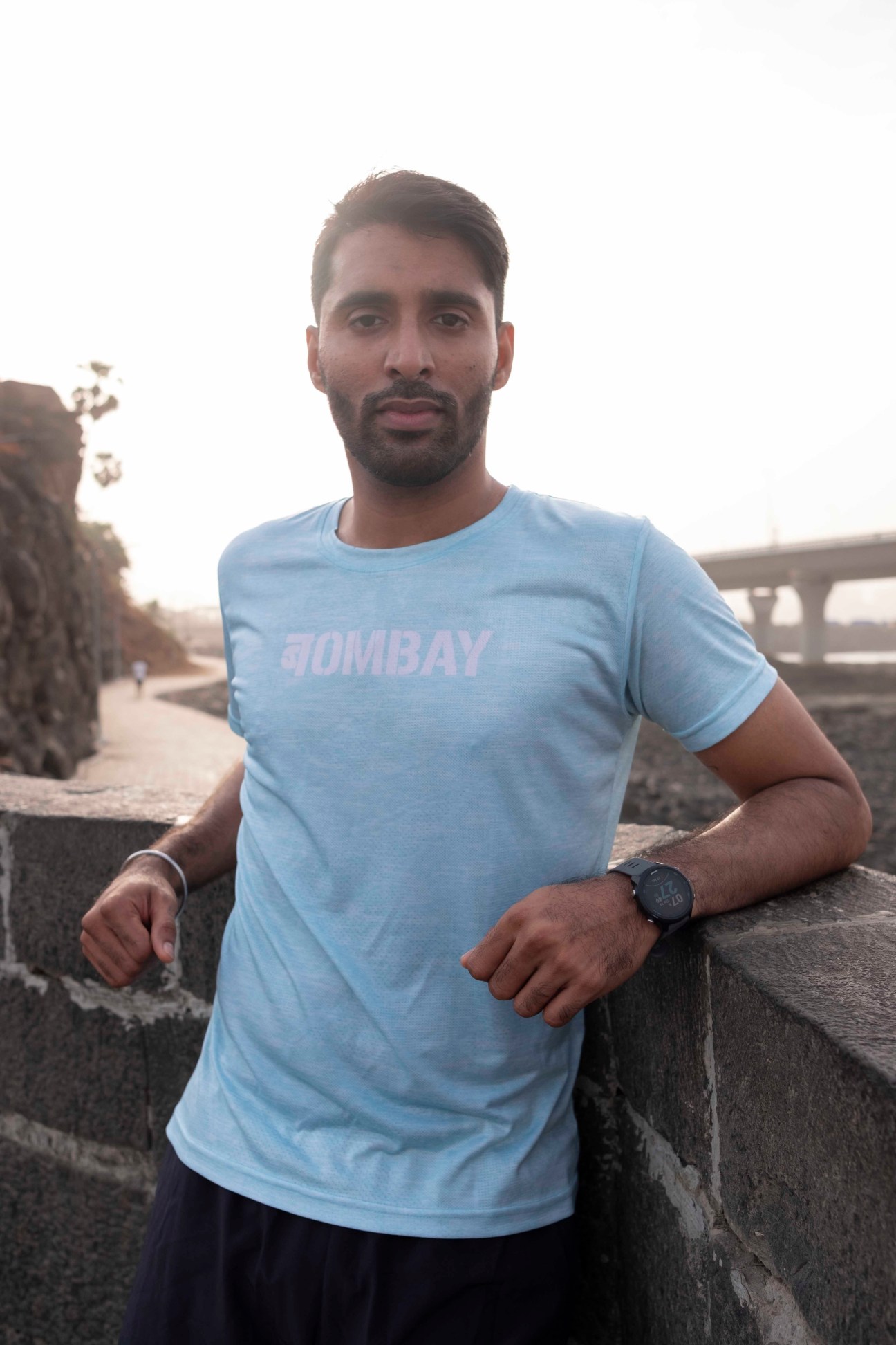 Man in light blue Bombay t-shirt leaning against concrete ledge outdoors