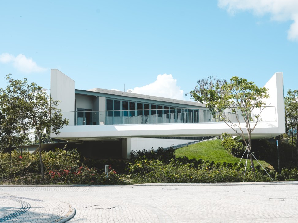Modern white concrete building with glass facade elevated on pillars above landscaped grounds