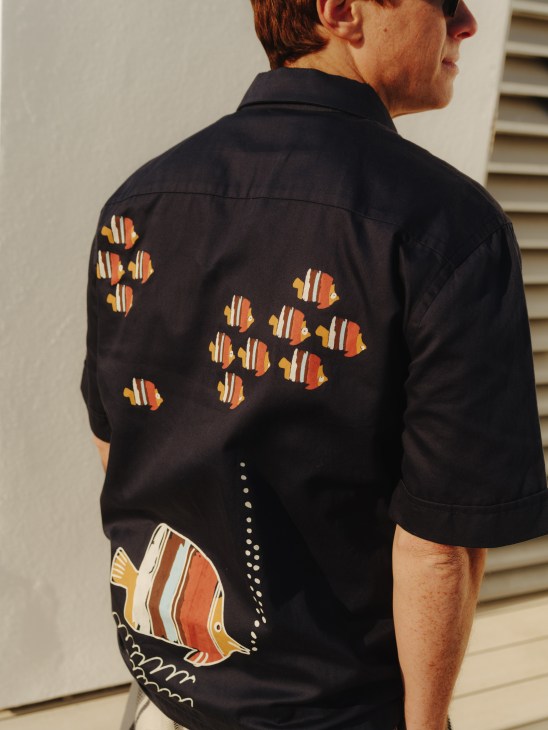 Man wearing black shirt with colorful tropical fish embroidery on back, showing detailed orange and white striped fish design