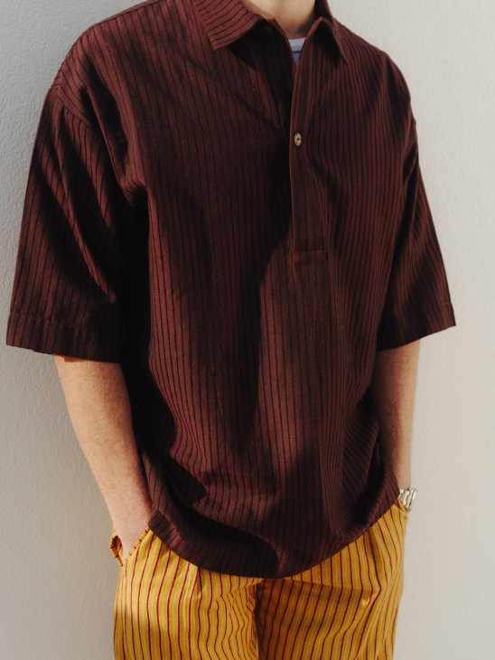 Person wearing burgundy ribbed short-sleeve shirt with yellow striped trousers against white wall