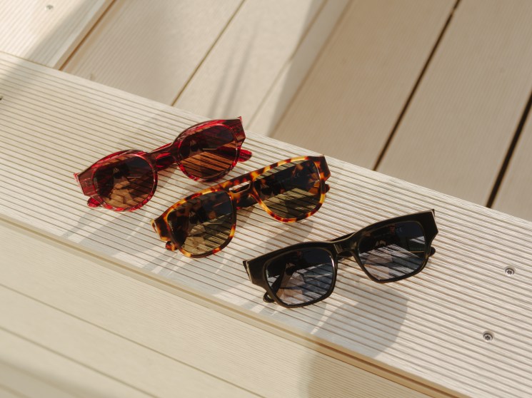 Three pairs of sunglasses in red, tortoiseshell, and black frames arranged on a wooden deck surface