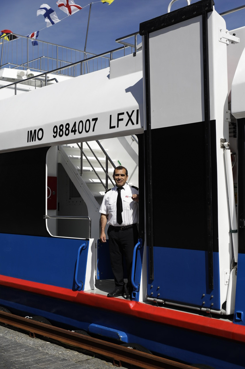 Ship crew member in white uniform standing in doorway of vessel IMO 9884007 LFXI with maritime flags above