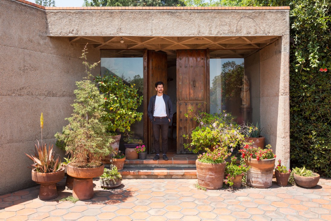 Man standing in doorway of Mexico City home surrounded by potted plants and flowering flowers on terrace.