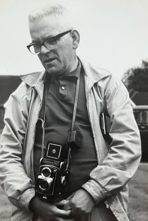 Young John Balsom as a child in casual clothing standing outdoors in the 1970s with glasses holding a twin-lens reflex camera, wearing a light jacket