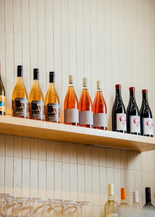 Wine bottles arranged on wooden shelf displaying orange, rosé and red wines with various labels
