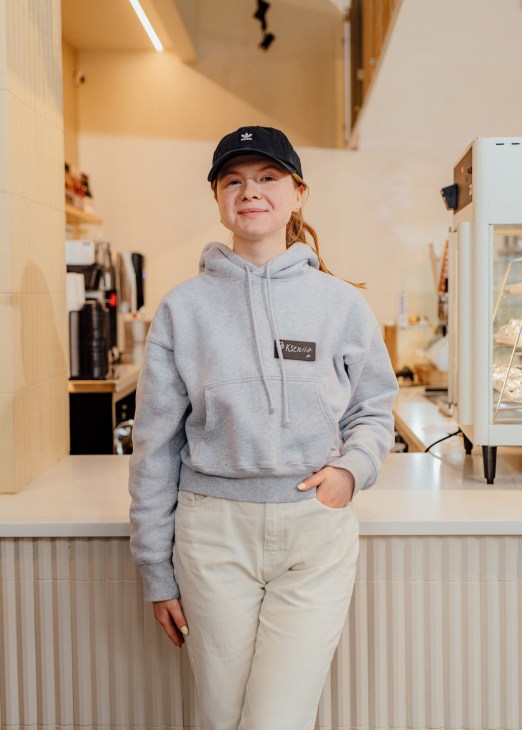 Young woman in gray hoodie and black cap smiling behind cafe counter with espresso machine