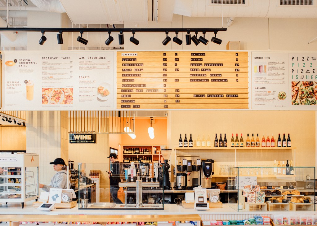 Modern cafe counter with menu boards, coffee equipment, wine display and staff member in bright interior