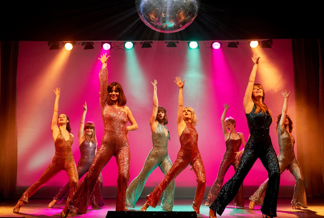 Eight women in sequined jumpsuits perform a synchronized dance routine on stage with colorful spotlights and disco ball