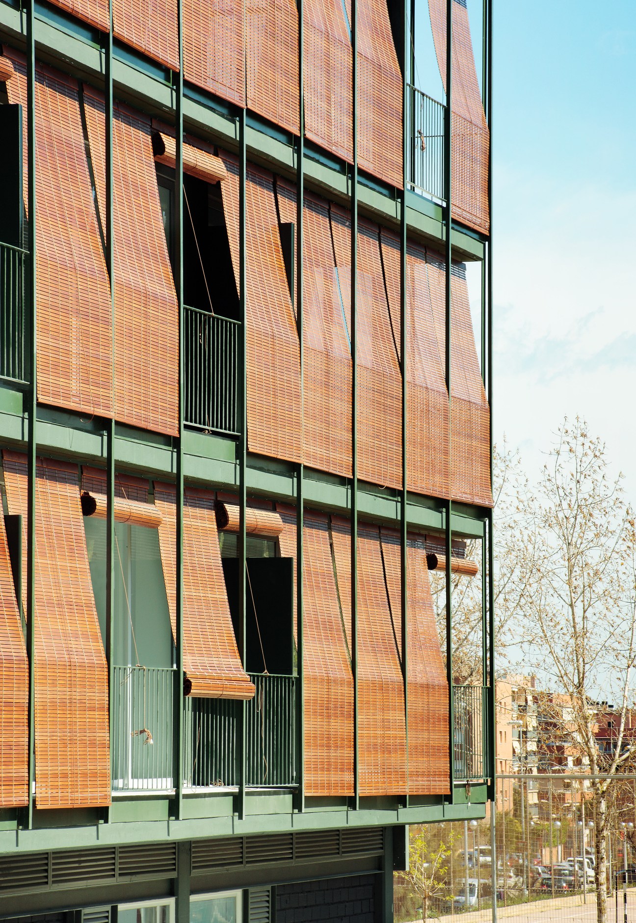 Modern brick apartment building with green metal framing and balconies overlooking bare trees.