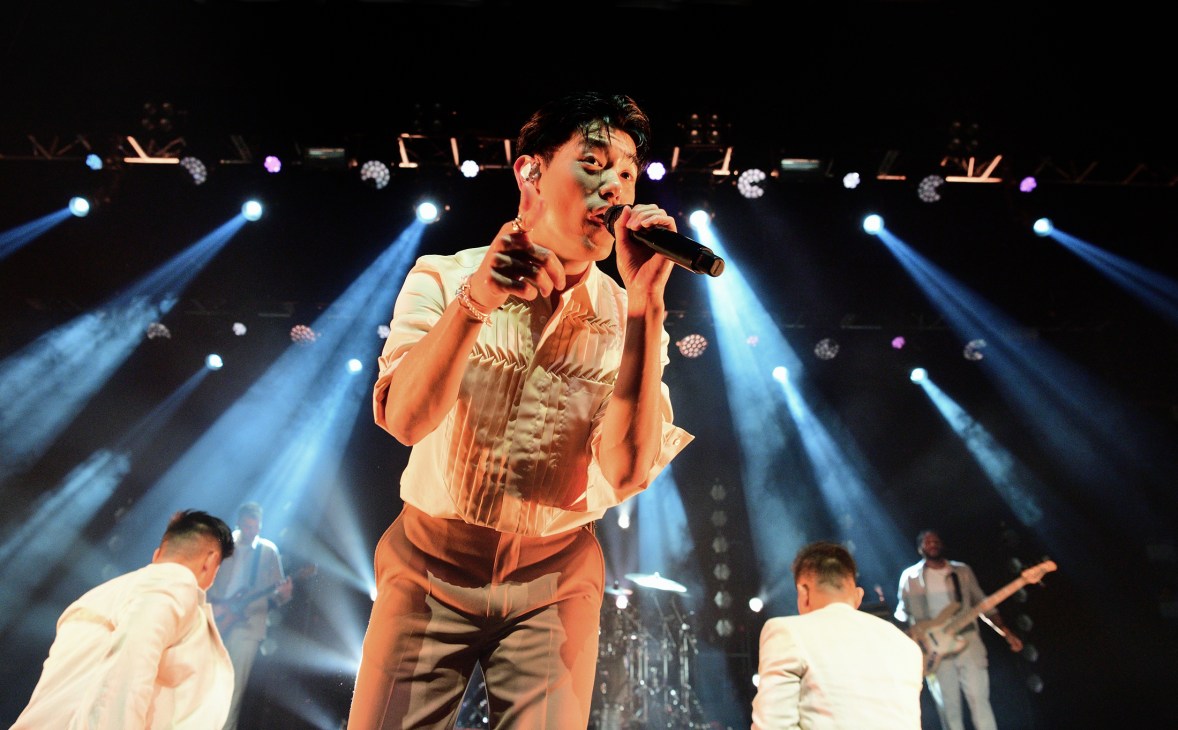 Eric Nam performing on stage with microphone, wearing white shirt with stage lighting in background