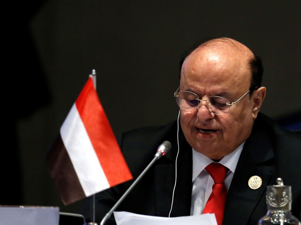 Man in suit and glasses speaking at microphone with red and white flag in background