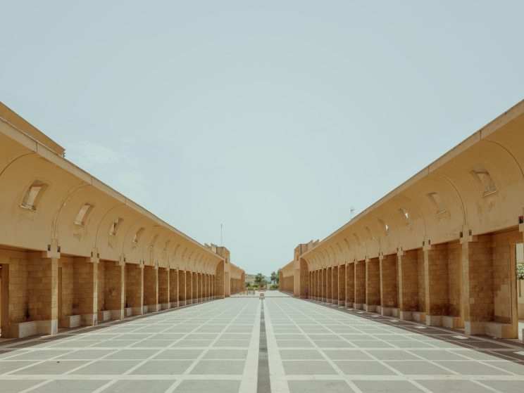 Symmetrical sand-colored stone colonnade with arched openings flanking a geometric paved courtyard under pale sky