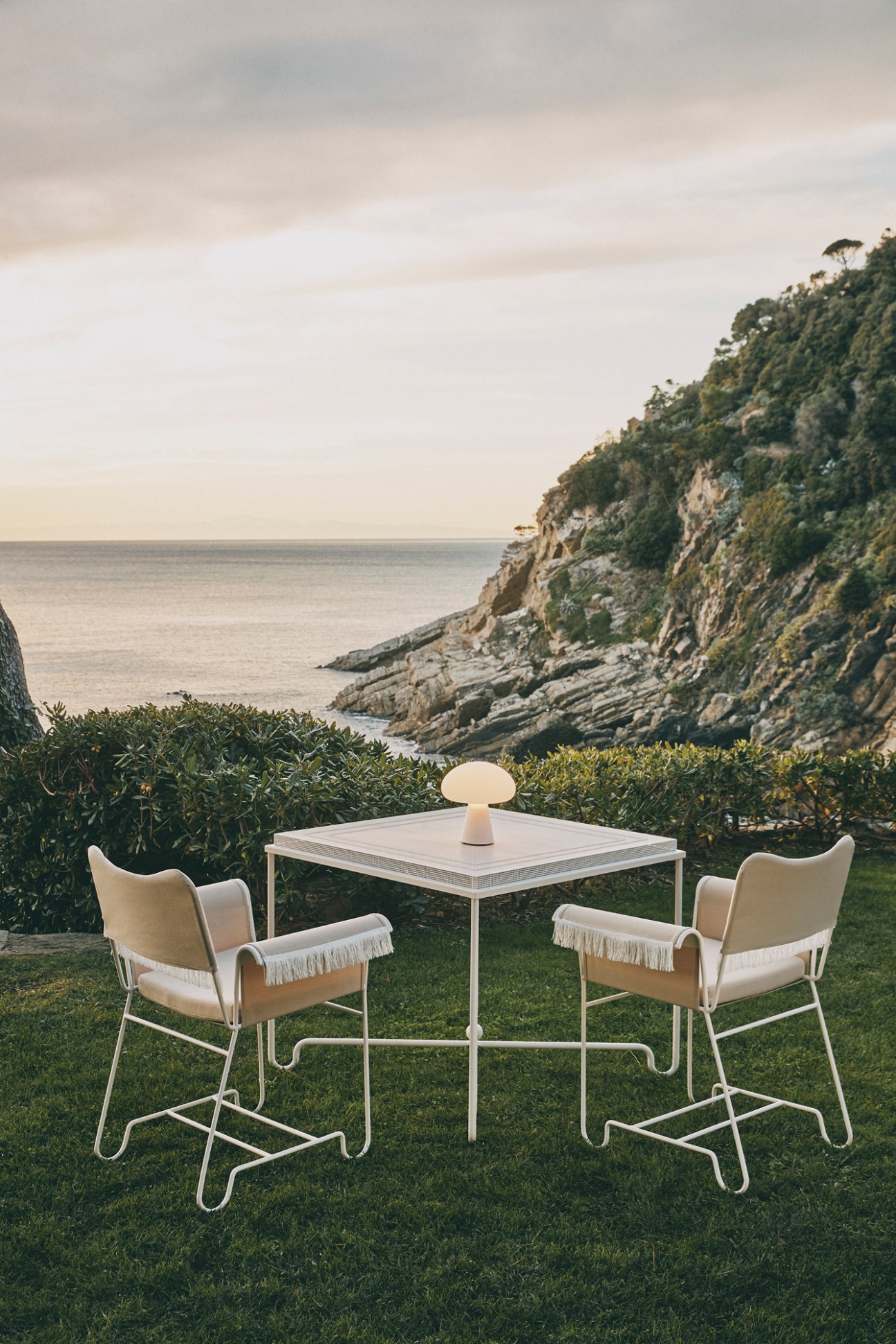Gubi Tropique Collection outdoor furniture set with cream canvas chairs and white table on grass overlooking ocean