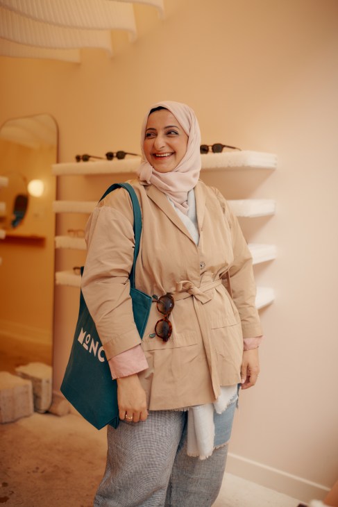 Woman wearing hijab and beige coat holding teal bag with Monc text, smiling in warm-lit interior space