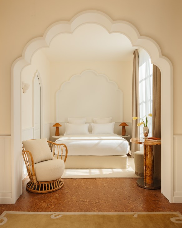 Hotel Lilou Chambre Mauresque scallopped archway opening into a double bed with white bedding and a scallopped white tall headboard floor-to-ceiling window with drapes ratan armchair with plush white cushions and marbled table brown cork patterned flooring with white carpet under bed and a rug