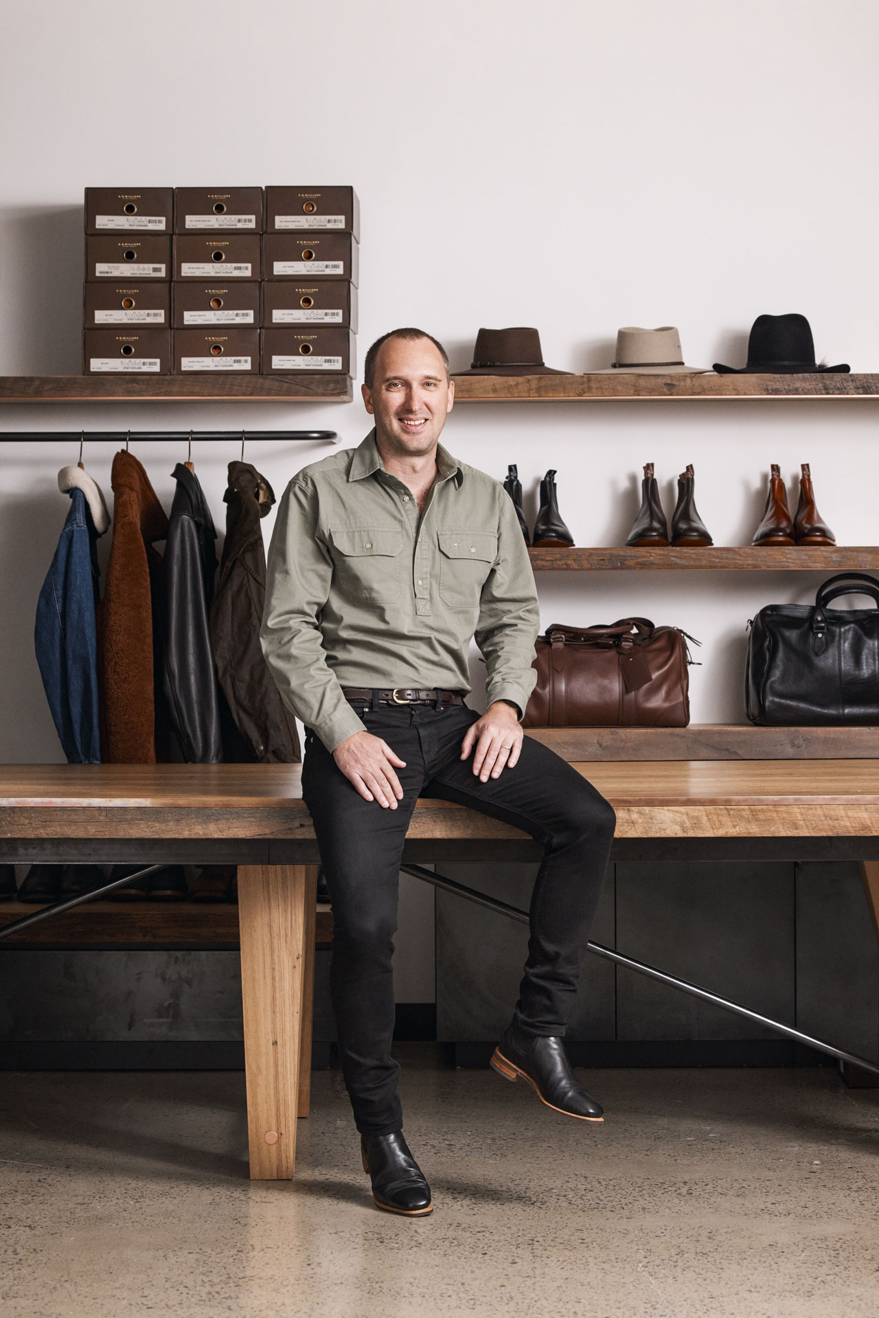 CEO Paul Grosmann sitting on wooden bench in retail store surrounded by leather goods, hats and shoe boxes