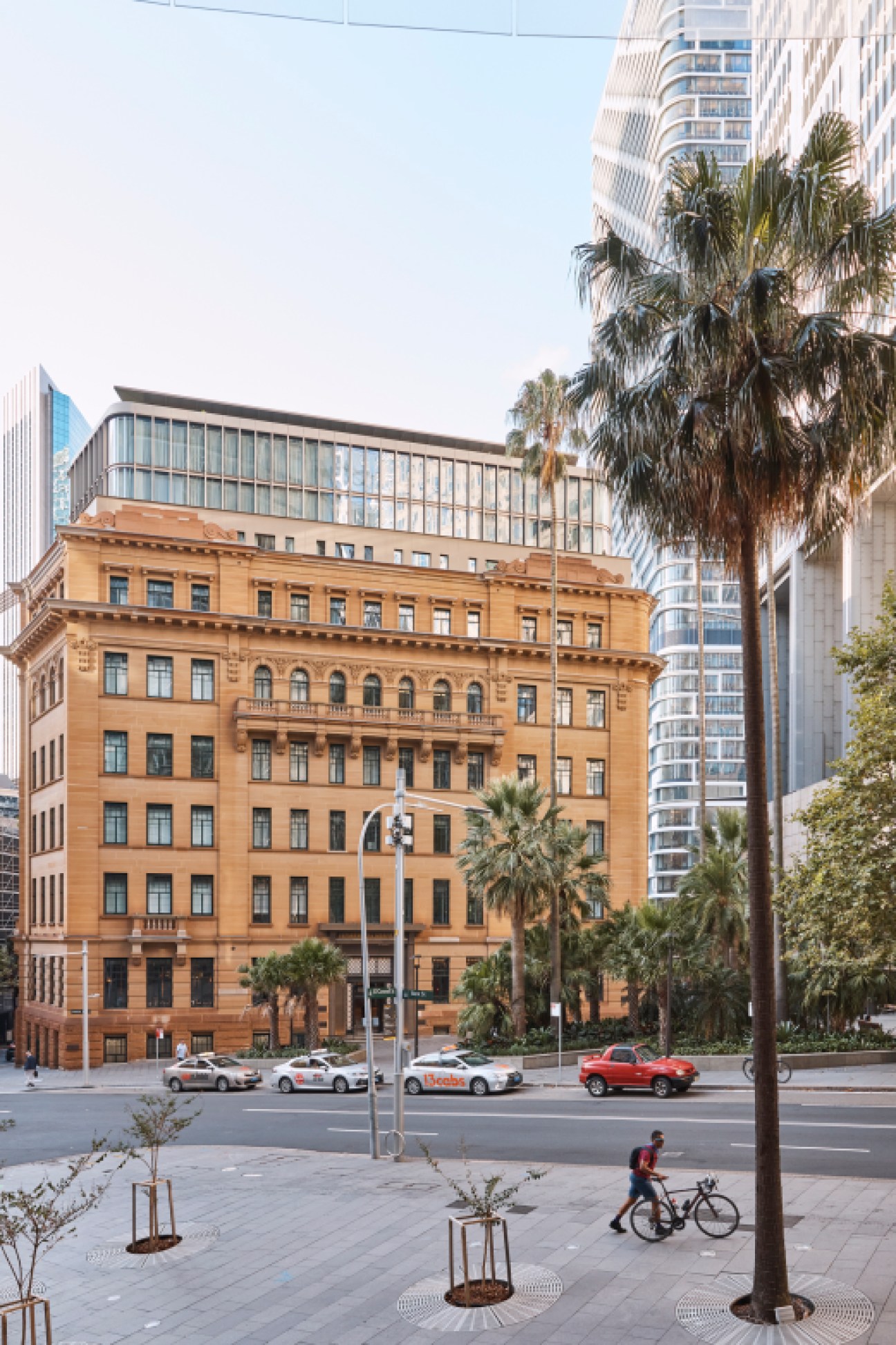 Historic sandstone building with classical architecture, modern glass addition on top, surrounded by palm trees and skyscrapers