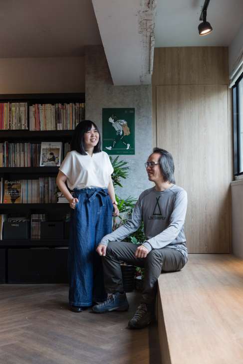Two people pose in a modern office space with bookshelves and plants, one standing and one crouching.