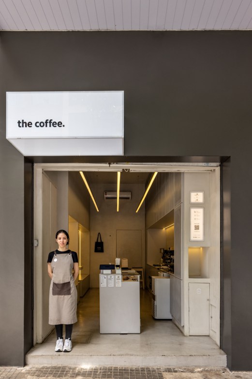 A female barista in an apron stands at the entrance of a small, minimalist coffee shop with modern lighting