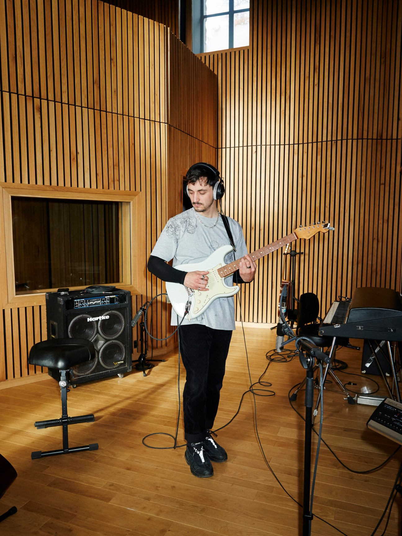 Musician playing electric guitar in a recording studio with wooden vertical slat walls and acoustic equipment.