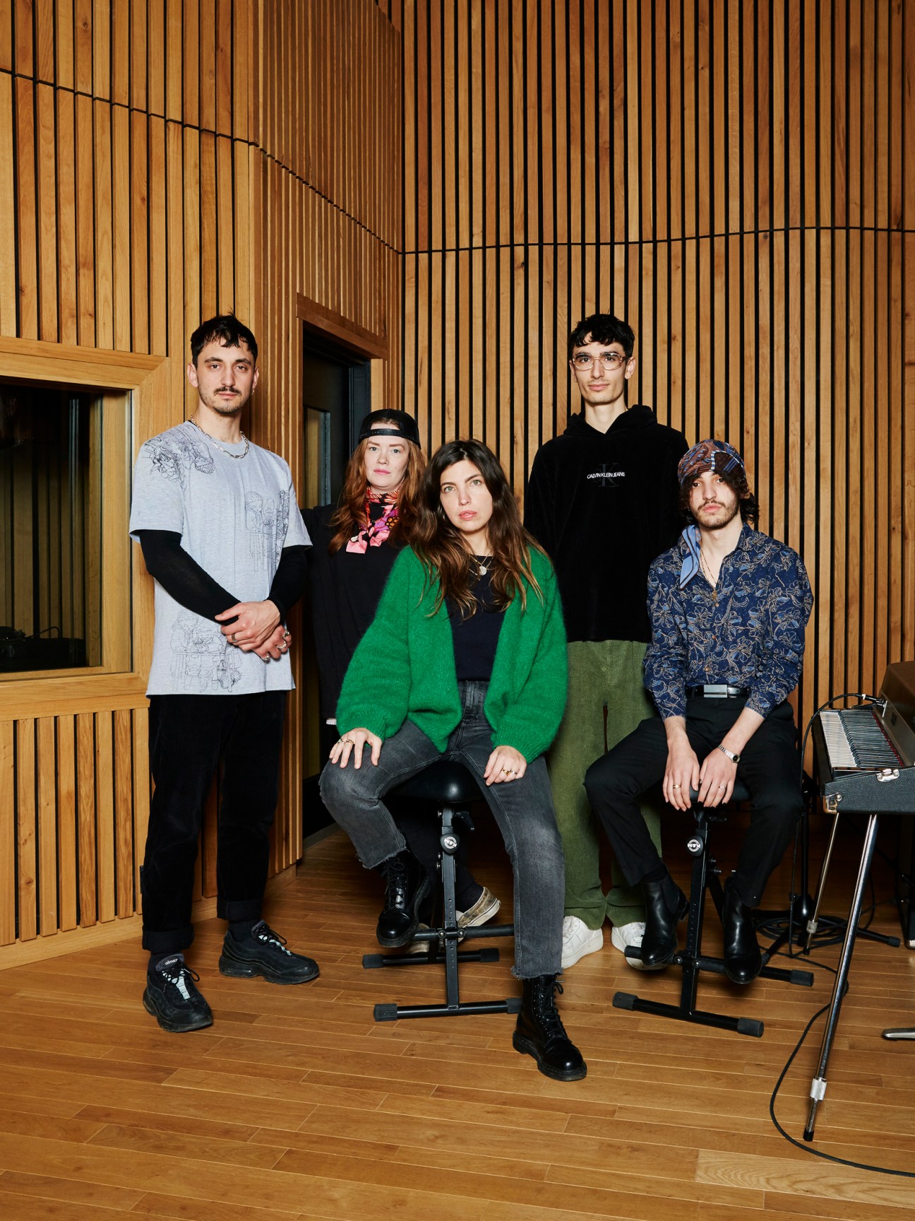 Five musicians posing together in a wooden-paneled studio room, with Clou in the centre wearing a green cardigan.