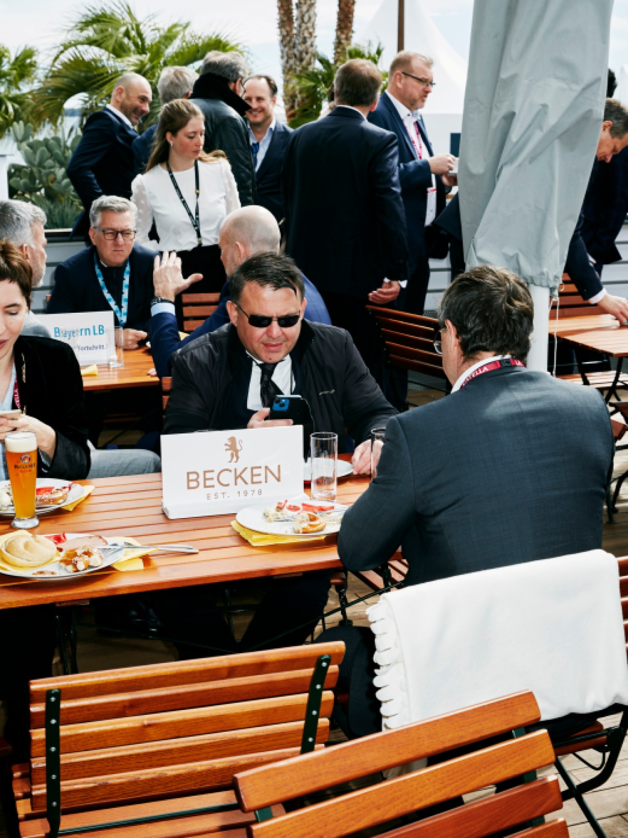 Business professionals in suits networking at outdoor tables during Mipim trade fair with palm trees in background
