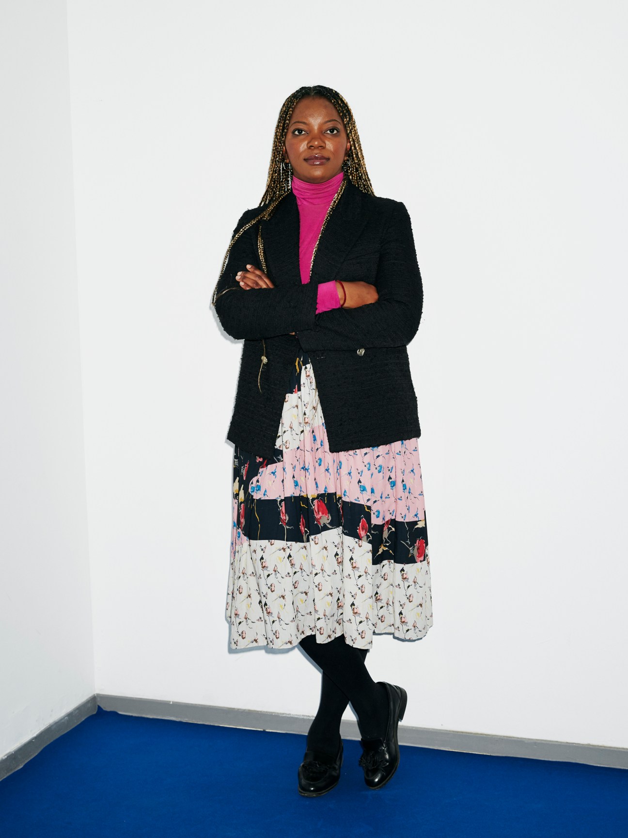 Professional woman in black blazer and bright pink turtleneck standing with arms crossed against white wall