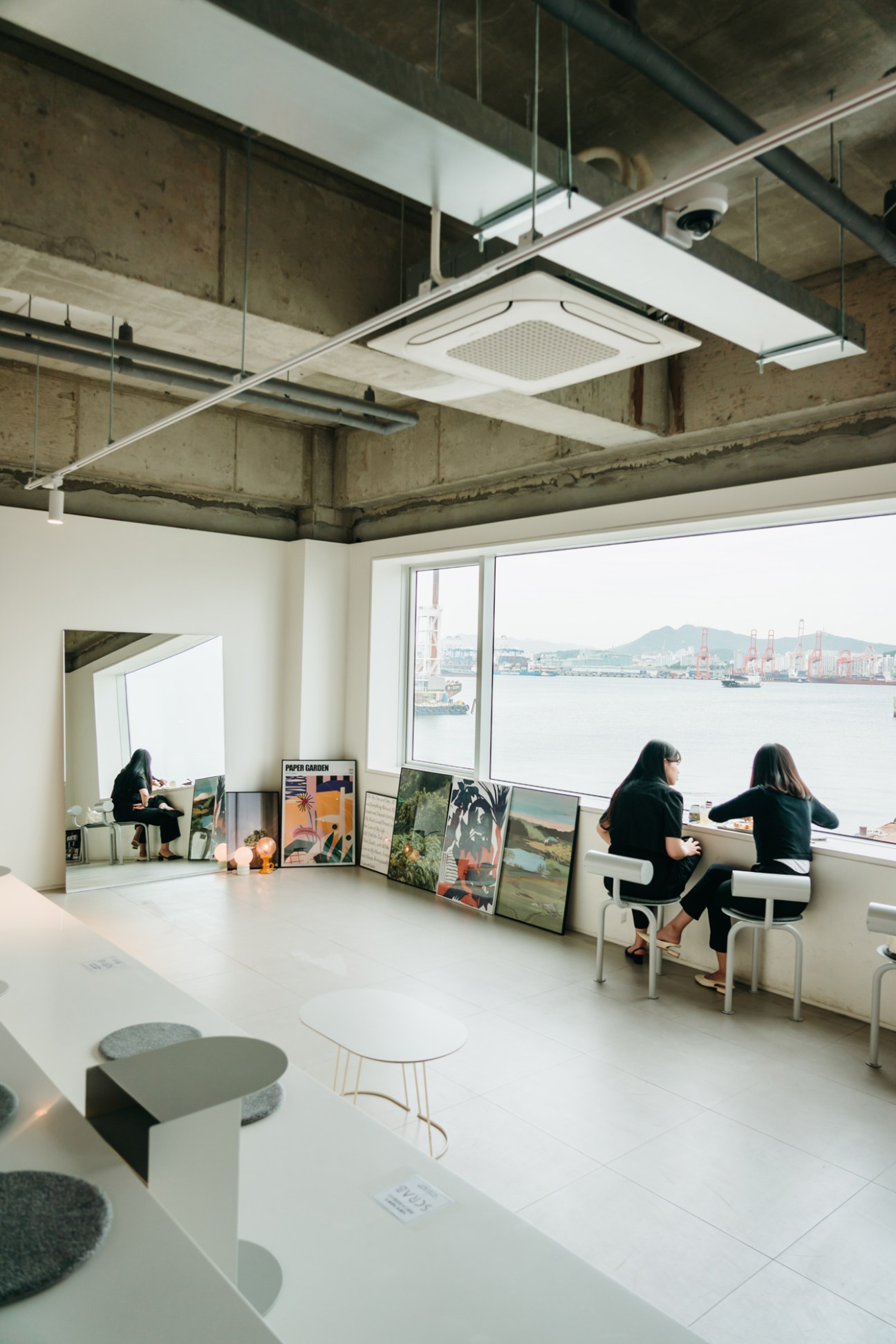 Modern office space with large windows overlooking water, two people working at desks with exposed concrete ceiling